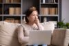 Eine ältere Frau sitzt auf dem Sofa mit Laptop auf dem Schoß und blickt nachdenklich zur Seite.