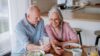 Ein älterer Mann und eine ältere Frau sitzen gemeinsam am Esstisch, essen und schauen auf ein Smartphone.
