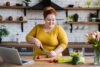 Eine junge, übergewichtige Frau bereitet in der Küche einen Salat zu. Vor Ihr auf der Arbeitsfläche steht ihr Laptop.